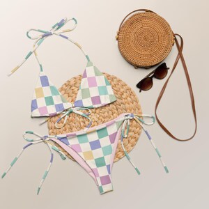 May include: A checkered bikini with a light blue, green, yellow, and purple pattern. The bikini top is a triangle style and the bottom is a tie-side style. The bikini is on a woven straw mat with a round woven straw bag and sunglasses.