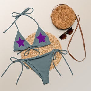 May include: A gray bikini with purple star accents on the top. The bikini is laid out on a woven straw mat with a straw bag and sunglasses.