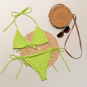 May include: A lime green bikini with a triangle top and a high-cut bottom. The bikini is on a woven straw placemat with a round straw bag and sunglasses.