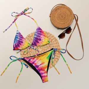 May include: A tie-dye bikini set with a triangle top and a high-cut bottom. The bikini is on a woven straw mat with a round straw bag and sunglasses.
