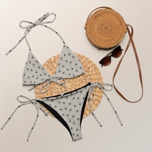 May include: A gray and black spider print bikini with a matching top and bottom. The bikini is on a woven straw mat with a round woven straw bag and sunglasses.