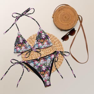 May include: A two-piece bikini set with a dark floral pattern featuring skulls and horror movie characters. The top and bottom have tie closures. A round woven bag and sunglasses are also in the image.