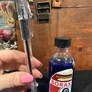 May include: A clear pen with a black ink cartridge next to a small blue glass bottle with a white label that reads "90% EFFECTIVE SELF DEFENSEMAX LEVIATHAN VILLAIN BORAX". The label also features a black and white illustration of a cartoon monster with a large mouth.