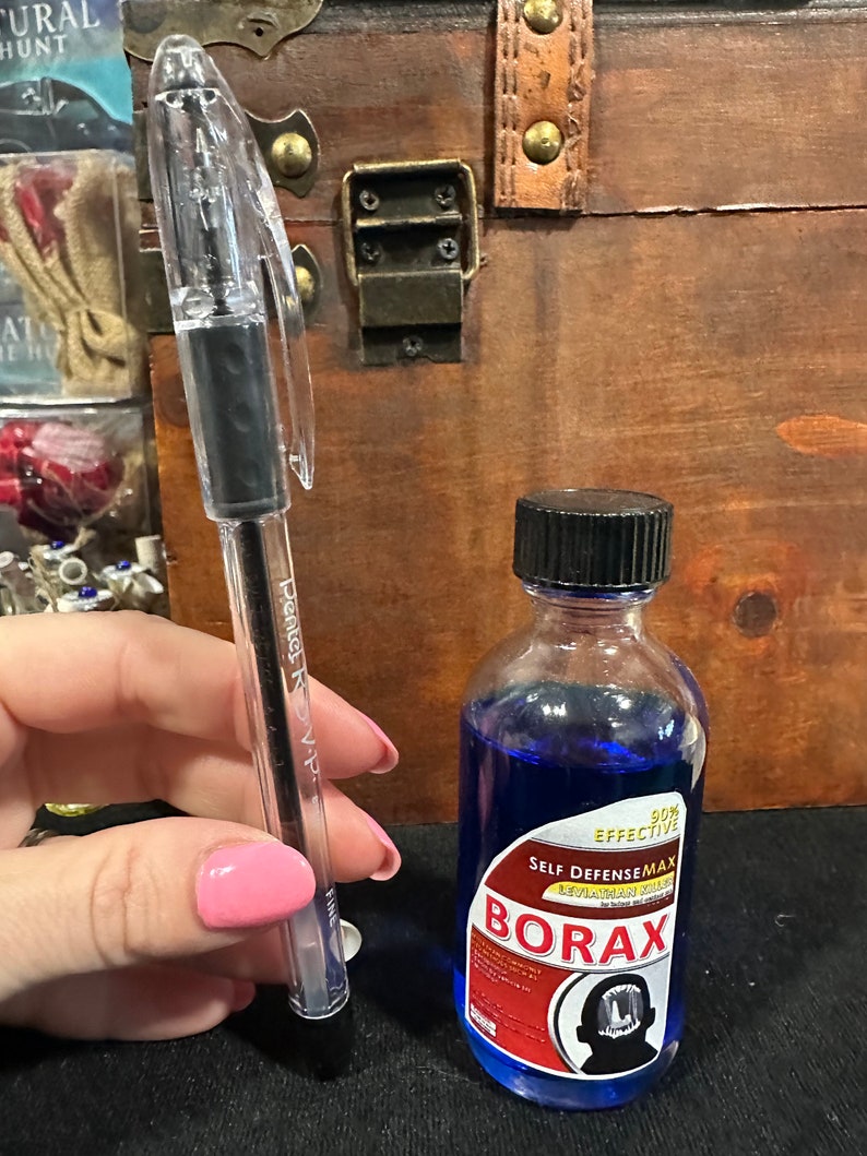 May include: A clear pen with a black ink cartridge next to a small blue glass bottle with a white label that reads "90% EFFECTIVE SELF DEFENSEMAX LEVIATHAN VILLAIN BORAX". The label also features a black and white illustration of a cartoon monster with a large mouth.
