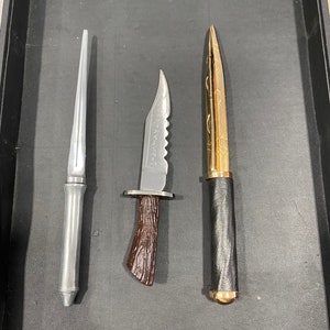 May include: Three different knives with different blade styles and handles. The knife on the left has a silver blade and handle. The knife in the middle has a silver blade with a brown wooden handle. The knife on the right has a gold blade with a black handle.