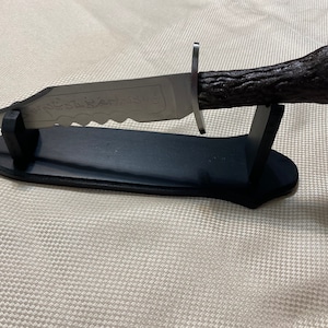 Może przedstawiać: Srebrny nóż z czarną drewnianą rękojeścią i ząbkowanym ostrzem. Nóż znajduje się na czarnym stojaku wystawowym. Ostrze ma napis "The Assassin's Creed".