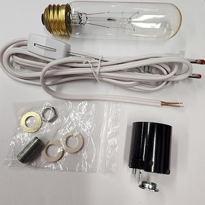 Puede incluir: Un conjunto de componentes para la fabricación de lámparas, que incluye una bombilla de vidrio transparente con base de latón, un portalámparas negro, un cable eléctrico blanco con interruptor y varias piezas metálicas.