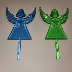 May include: Two plastic angel ornaments, one blue and one green, with wings outstretched and a stick for inserting into a Christmas tree or other festive arrangement.