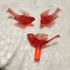 May include: Three red plastic bird ornaments for a Christmas tree. The birds have clear wings and are in a flying position.