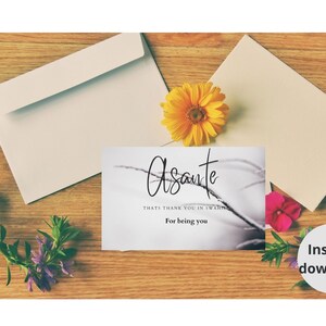 May include: A white card with black text that reads "Asante" which means "thank you" in Swahili. The card also says "That's thank you in Swahili" and "For being you". The card is on a wooden surface with flowers.