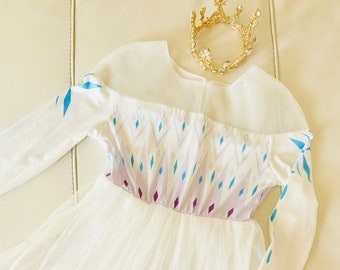 May include: A white and blue princess dress with long sleeves and a tulle skirt. The bodice features a diamond pattern in blue and purple. A gold crown with clear gems sits nearby. The dress has a flowing, elegant design.