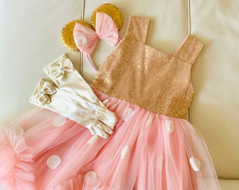 May include: A pink tulle dress with white polka dots and a gold sequin bodice. The dress has a ruffled hem and comes with a pair of white gloves and a gold headband with pink bow.