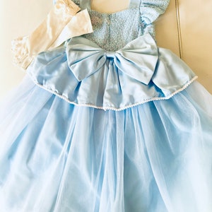 K&ouml;nnte beinhalten: Ein hellblaues T&uuml;llkleid mit einer wei&szlig;en Schleife aus Satin und wei&szlig;en Spitzenhandschuhen. Das Kleid hat ein Paillettenoberteil und einen Faltenrock.