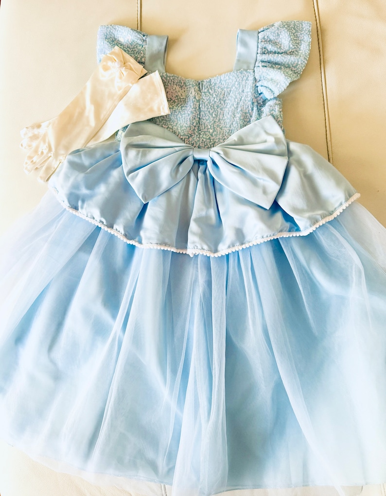 K&ouml;nnte beinhalten: Ein hellblaues T&uuml;llkleid mit einer wei&szlig;en Schleife aus Satin und wei&szlig;en Spitzenhandschuhen. Das Kleid hat ein Paillettenoberteil und einen Faltenrock.