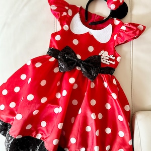 May include: Red polka dot dress with black sequin trim and a black bow. The dress has a white collar and a white embroidered patch that says "Love that Good ol' fashioned Nature". The dress is paired with a black headband with red polka dot ears and a red bow.