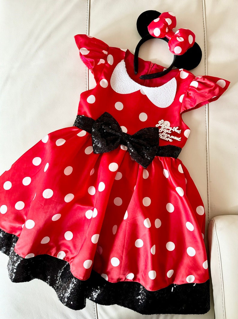 May include: Red polka dot dress with black sequin trim and a black bow. The dress has a white collar and a white embroidered patch that says "Love that Good ol' fashioned Nature". The dress is paired with a black headband with red polka dot ears and a red bow.