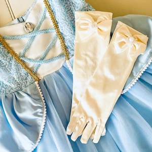 K&ouml;nnte beinhalten: Ein hellblaues T&uuml;llkleid mit einem wei&szlig;en Satin-Oberteil und goldenen Verzierungen. Das Kleid hat einen wei&szlig;en Perlenbesatz am Saum. Zum Kleid geh&ouml;rt ein Paar wei&szlig;e Satinhandschuhe mit einer wei&szlig;en Satin Schleife und Perlenverzierung.