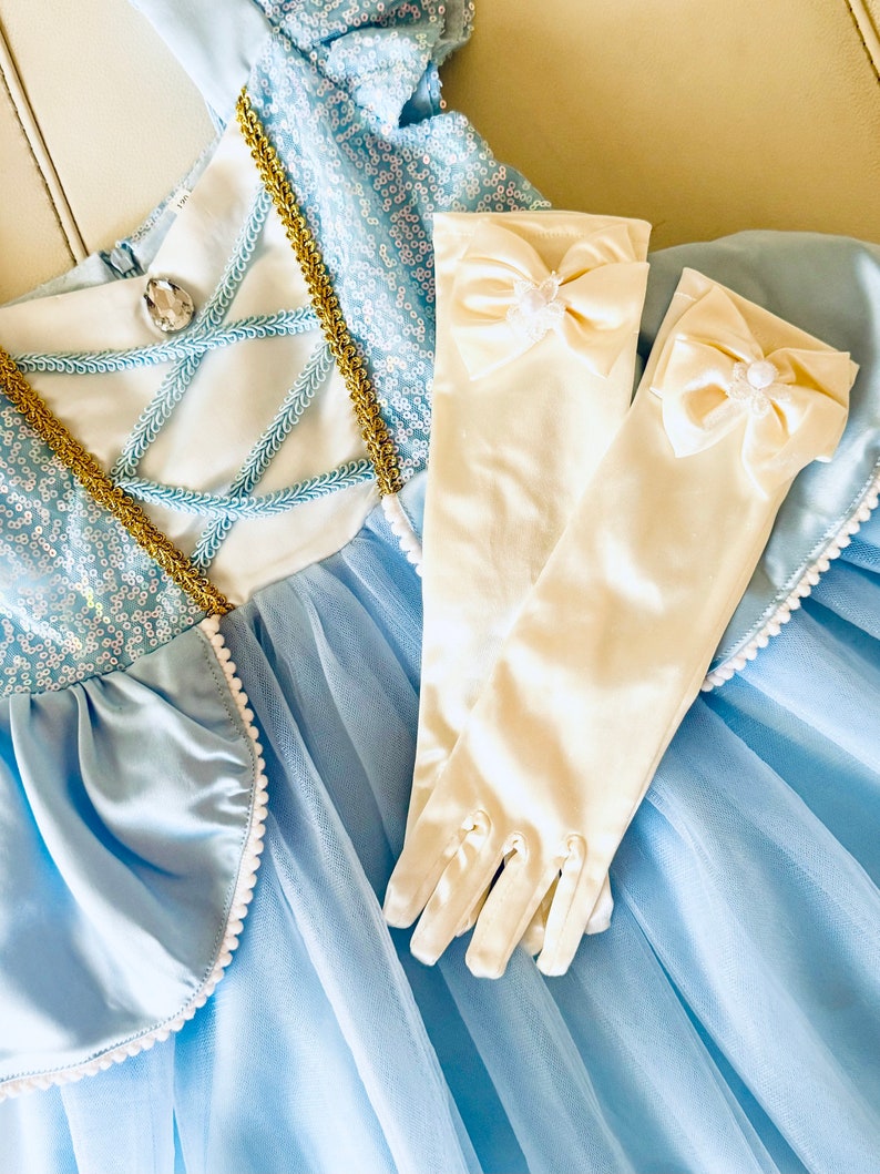 K&ouml;nnte beinhalten: Ein hellblaues T&uuml;llkleid mit einem wei&szlig;en Satin-Oberteil und goldenen Verzierungen. Das Kleid hat einen wei&szlig;en Perlenbesatz am Saum. Zum Kleid geh&ouml;rt ein Paar wei&szlig;e Satinhandschuhe mit einer wei&szlig;en Satin Schleife und Perlenverzierung.