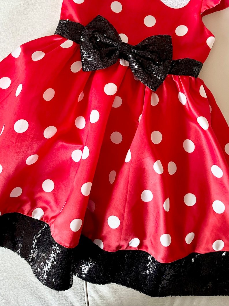 May include: Red dress with white polka dots and a black sequin bow. The dress has a black sequin trim around the bottom.