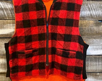 Könnte beinhalten: Eine Vintage-Weste ohne Ärmel in einem rot-schwarzen Büffel-Plaid-Muster. Die Weste hat einen schwarzen Reißverschluss, schwarze Paspeln und zwei Vordertaschen. Die Innenseite ist orange. Die Weste hängt an einem Holzbügel vor einem Holzgrund.