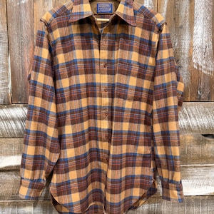 May include: A long-sleeved button-down shirt featuring a plaid pattern in shades of tan, brown, and blue. The shirt has a pointed collar, a single chest pocket, and a blue Pendleton brand label sewn into the inner neck area of the garment.