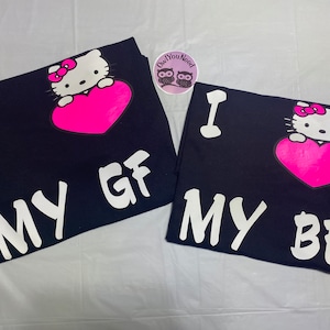 May include: Two navy blue t-shirts with a Hello Kitty graphic holding a pink heart. One shirt says "I MY GF" and the other says "I MY BF" in white block letters. A round sticker with owls is in the background.