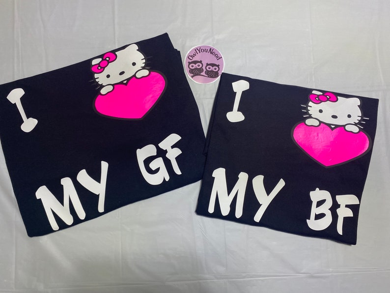 May include: Two navy blue t-shirts with a Hello Kitty graphic holding a pink heart. One shirt says "I MY GF" and the other says "I MY BF" in white block letters. A round sticker with owls is in the background.