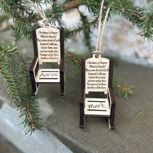 May include: Two wooden chair ornaments with the text "Christmas in Heaven" and a poem. One ornament is labeled "Aaron" and the other "Mark". The ornaments are hanging from twine and are displayed against a backdrop of evergreen branches.