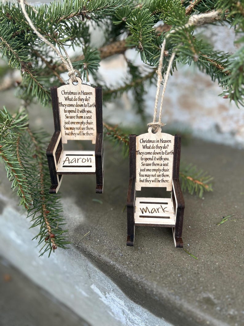 May include: Two wooden chair ornaments with the text "Christmas in Heaven" and a poem. One ornament is labeled "Aaron" and the other "Mark". The ornaments are hanging from twine and are displayed against a backdrop of evergreen branches.