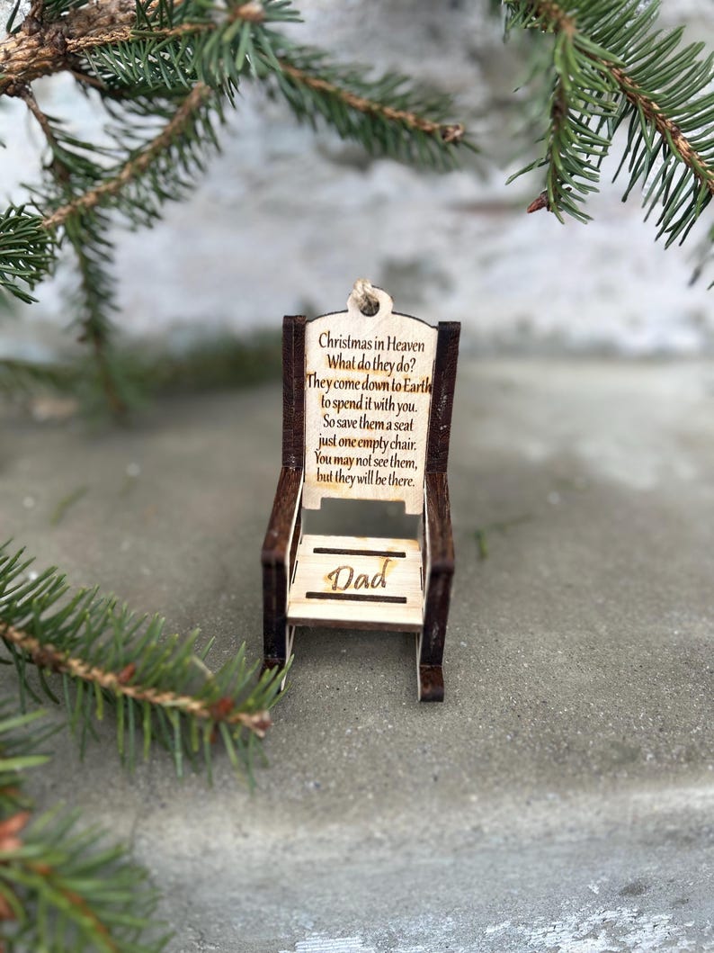 May include: A wooden chair ornament with the text "Christmas in Heaven" and a poem about remembering loved ones. The word "Dad" is inscribed on the chair. The ornament is brown and beige, with a natural wood grain finish.
