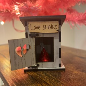 May include: A small, wooden outhouse with a hinged door and a sign that reads "Love stinks." The door has a heart-shaped cutout. Inside is a red foil-wrapped chocolate. The outhouse is on a wooden surface.