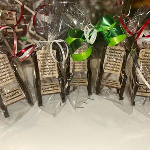 May include: Small wooden chair ornaments, each wrapped in clear cellophane and tied with a ribbon. Each chair has a small sign with a poem and a name: Daddy, Grandpap, Nana, and Pap. The chairs are brown with a light wood sign.