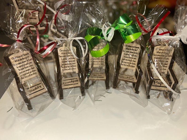 May include: Small wooden chair ornaments, each wrapped in clear cellophane and tied with a ribbon. Each chair has a small sign with a poem and a name: Daddy, Grandpap, Nana, and Pap. The chairs are brown with a light wood sign.