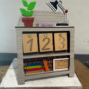 Könnte beinhalten: Ein graues Bücherregal aus Holz mit drei nummerierten Schubladen. Oben stehen eine grüne Pflanze in einem Glitzertopf und eine silberne Schreibtischlampe. Das untere Regal zeigt bunte Bücher und ein Schild mit der Aufschrift "Just One more Chapter."
