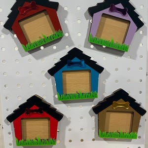 May include: A collection of colorful dog house-shaped picture frames. Each frame features a different color scheme, including red, purple, blue, and brown, with a black roof and a bone-shaped accent. Green grass decorates the bottom.