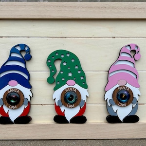 Puede incluir: Tres decoraciones de madera de Santa Cam en un marco de madera. Cada gnomo tiene un sombrero de diferente color: rayas azules y blancas, verde con estrellas blancas y rayas rosas y blancas. Cada uno tiene una lente de cámara y las palabras "SANTA CAM".