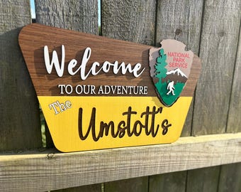Puede incluir: Letrero de madera con las palabras "Welcome to our Adventure" y el apellido "Umstott's". Presenta un emblema del Servicio de Parques Nacionales. El letrero tiene una combinación de colores marrón, amarillo y blanco.