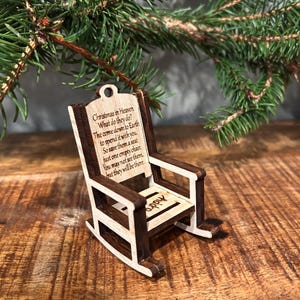 May include: A miniature wooden rocking chair ornament with a light wood frame and dark brown accents. The chair has the text "Christmas in Heaven" and a poem. The word "appy" is also visible on the seat. A Christmas tree branch is in the background.