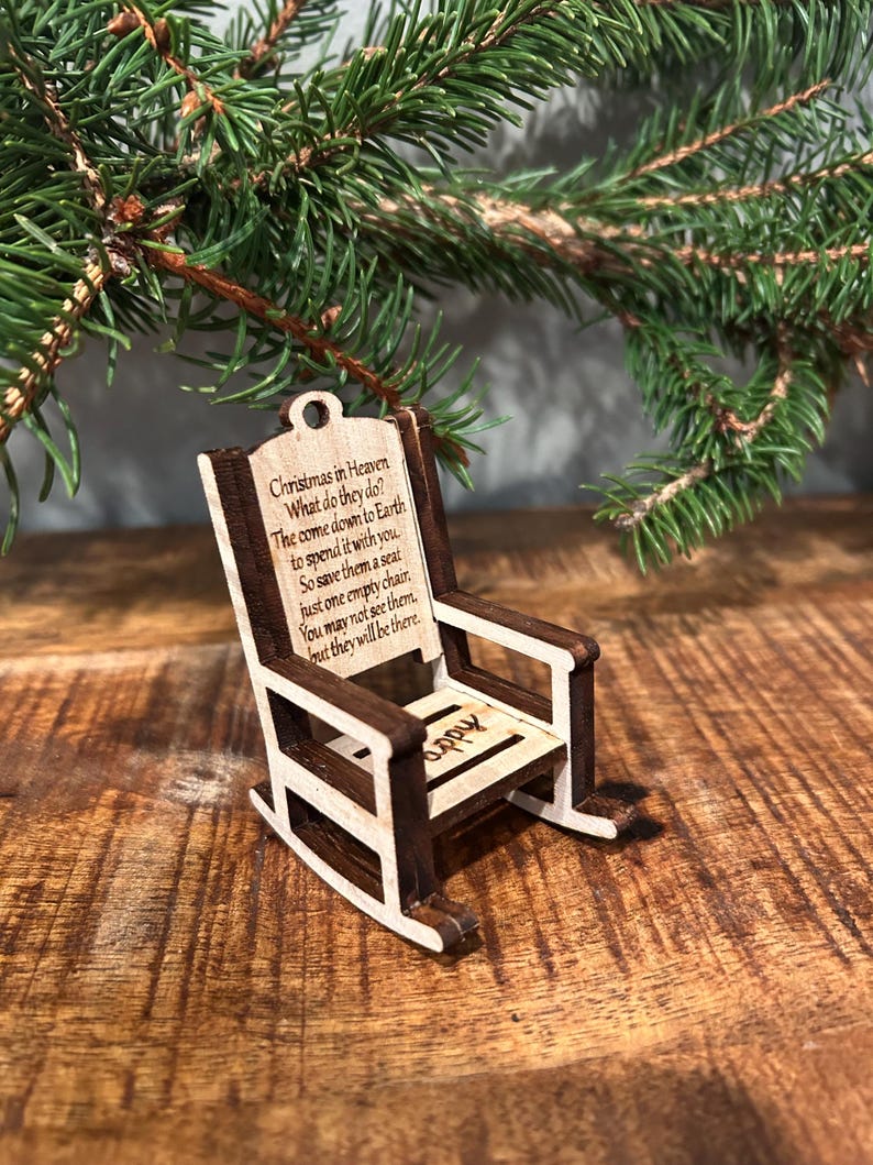 May include: A miniature wooden rocking chair ornament with a light wood frame and dark brown accents. The chair has the text "Christmas in Heaven" and a poem. The word "appy" is also visible on the seat. A Christmas tree branch is in the background.