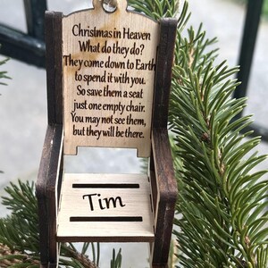 May include: A wooden chair ornament with the text "Christmas in Heaven" and a poem. The chair is light brown with dark brown trim and the name "Tim" is engraved on the seat. The ornament hangs from a natural twine string.