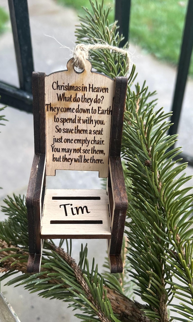 May include: A wooden chair ornament with the text "Christmas in Heaven" and a poem. The chair is light brown with dark brown trim and the name "Tim" is engraved on the seat. The ornament hangs from a natural twine string.