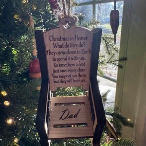 May include: A wooden chair-shaped Christmas ornament with the text "Christmas in Heaven" and "Dad". The ornament is hanging on a Christmas tree. The text on the ornament reads: "They come down to Earth to spend it with you. So save them a seat just one empty chair."
