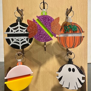 May include: Five circular acrylic keychains featuring Halloween designs: a black spiderweb, a purple glitter candy, an orange pumpkin, a yellow and orange candy corn, and a white ghost on a black glitter background, all attached to metal rings.