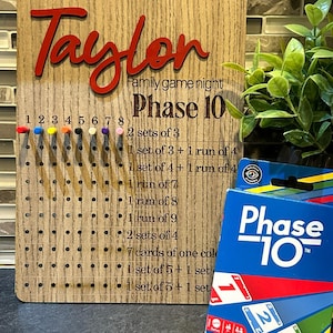 Könnte beinhalten: Ein Holz-Phase 10-Spielbrett mit dem Namen "Taylor" in roter Schrift. Das Brett hat Löcher für Stifte und eine Liste der Spielziele. Eine Schachtel mit Phase 10-Karten befindet sich neben dem Brett.
