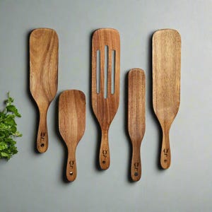 May include: Five wooden kitchen utensils, including spatulas and a slotted turner. The utensils are made of light brown wood with a natural grain pattern. Each has a handle with a small circular hole and a logo.