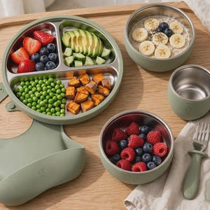 May include: A sage green silicone bib, a three-section stainless steel plate with strawberries, blueberries, avocado, cucumber, peas, and sweet potatoes, two stainless steel bowls with oatmeal and raspberries, a mug, a fork, and a spoon.