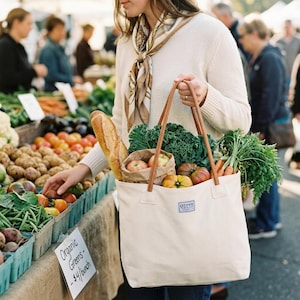 May include: A cream canvas tote bag with brown leather handles, filled with fresh vegetables and fruit. The bag contains a baguette, apples, tomatoes, carrots, and leafy greens. The bag has a label that reads "green essentials".