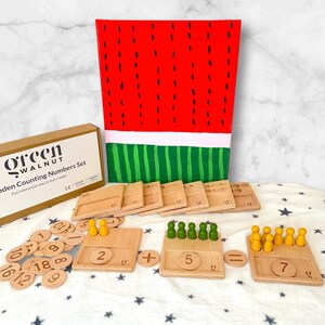 Puede incluir: Un juego de números de conteo de madera con un fondo temático de sandía. El juego incluye fichas de números de madera, tableros de conteo con clavijas y discos de madera. La caja dice "Green Walnut Wooden Counting Numbers Set".