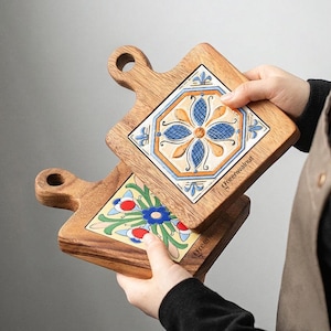 May include: Two wooden cutting boards with decorative ceramic tile inserts. The tiles feature floral patterns in blue, orange, and green. Each board has a handle and the text "Greenwalnut" is visible on the tile.