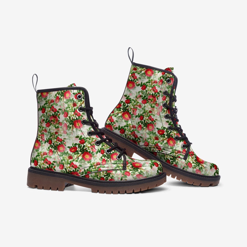 May include: A pair of combat boots with a floral pattern. The boots feature a white background with red roses and green leaves. They have black laces, black trim, and brown soles. The boots are a stylish footwear option.